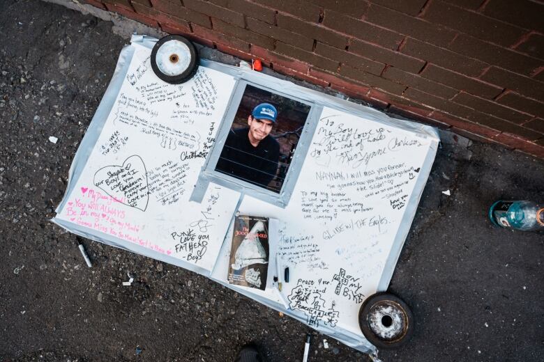 A whiteboard held down with two small wheels. It has a picture of a man in a blue hat and black shirt staring at the camera. The picture is surrounded by scrawled remembrances and messages.