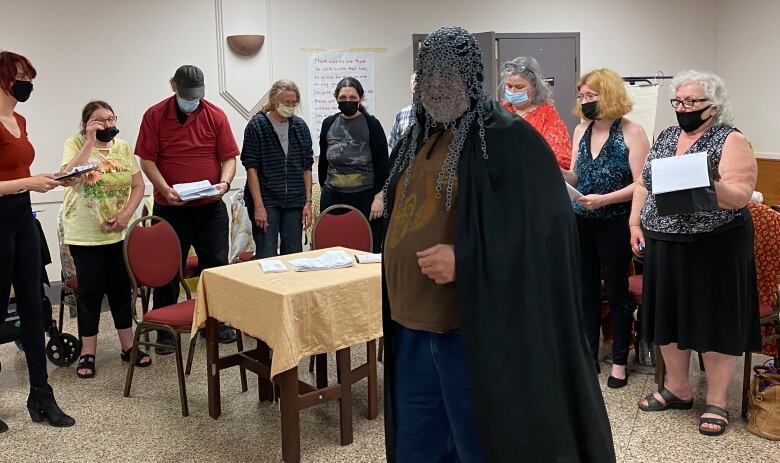 A man wearing a chainmail hood and robes stands in front of a group of people.