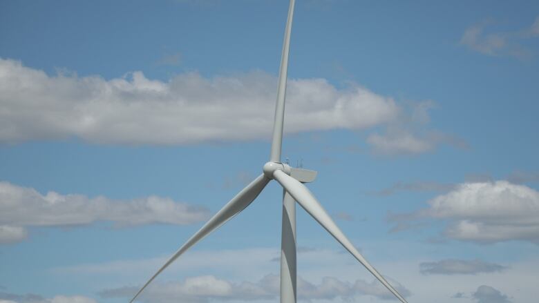 The top of a wind turbine is seen with blue sky and clouds in the background.