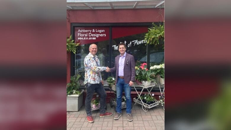 Two men shake hands in front of a florist's shop. One man has a speckled shirt and the other has a coat on.