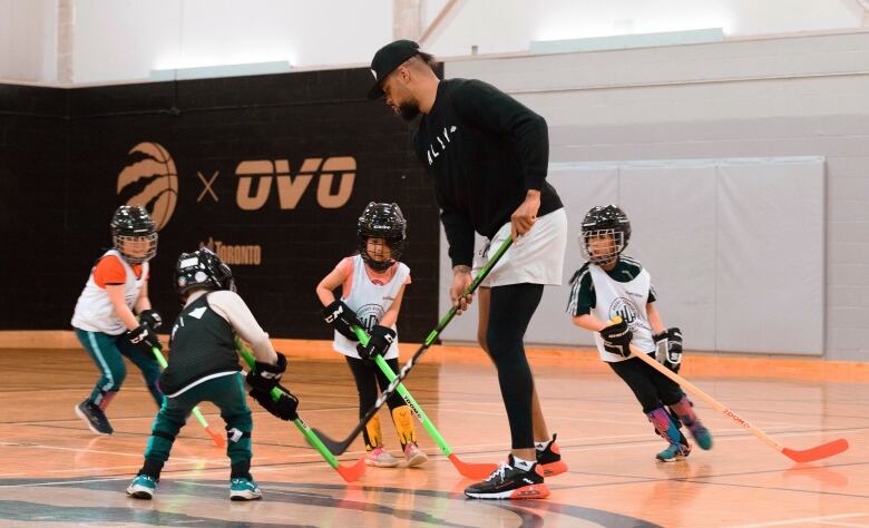 A man wearing a black sweater and black cap plays ball hockey in an indoor gym with young children wearing black helmets.