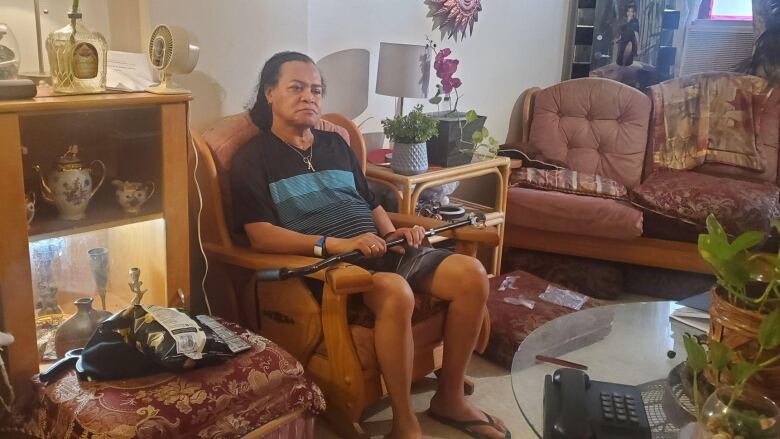 A man sits in an armchair with a cane across his knees in a small apartment.