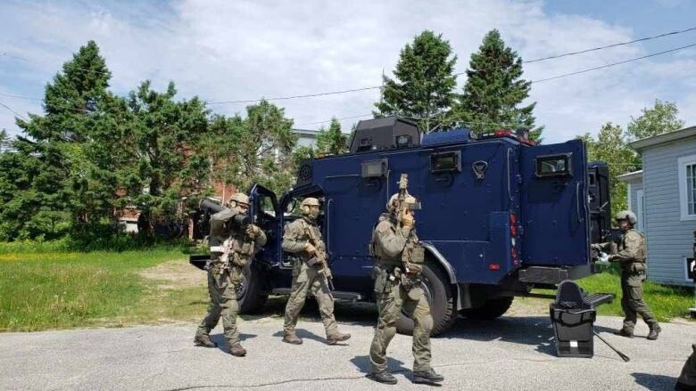 Four heavily armed police officers in camouflage gear are near a dark blue truck.