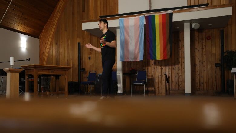 A man stands at the front of a church, talking and gesticulating. He is standing in front of two Pride flags and wearing a Pride T-shirt with a rainbow feather design.