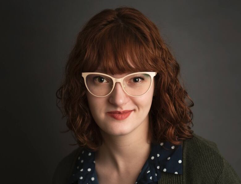 A white person in a blue polka dotted shirt and a green cardigan smiles at the camera. They are wearing white cat-eye glasses and are standing against a grey backdrop.