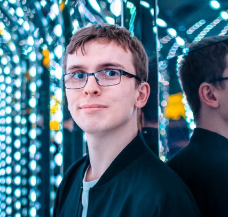 Liam is looking at the camera from an archway of mirrors and lights, like those found in a fairground. His back to the wall/mirror, with his hearing aid and glasses visible. He is wearing a black zip-up jacket and white top.