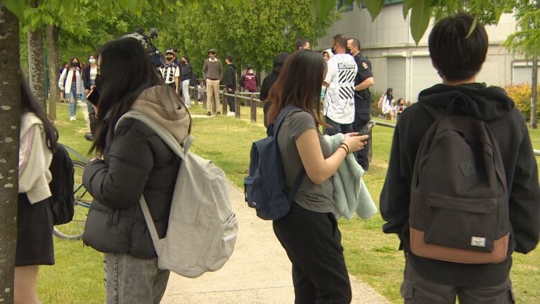 Students gather outside Killarney Secondary school in East Vancouver following a lockdown on Thursday, June 16, 2022. The backs of three students standing on a sidewalk beneath a leafy green tree can be seen in the foreground. 