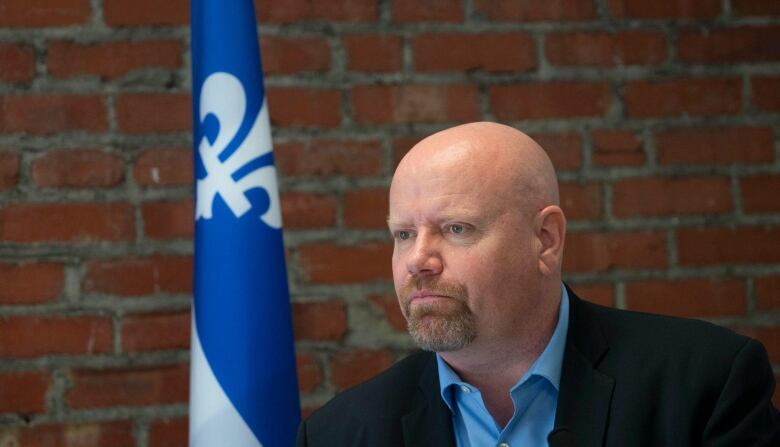 A serious-looking man sits in front of a brick wall, with a Quebec flag draped to his right.