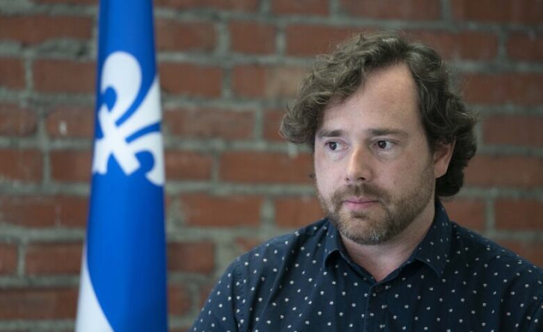 A young man sits in front of a brick wall, with a Quebec flag draped at his side.