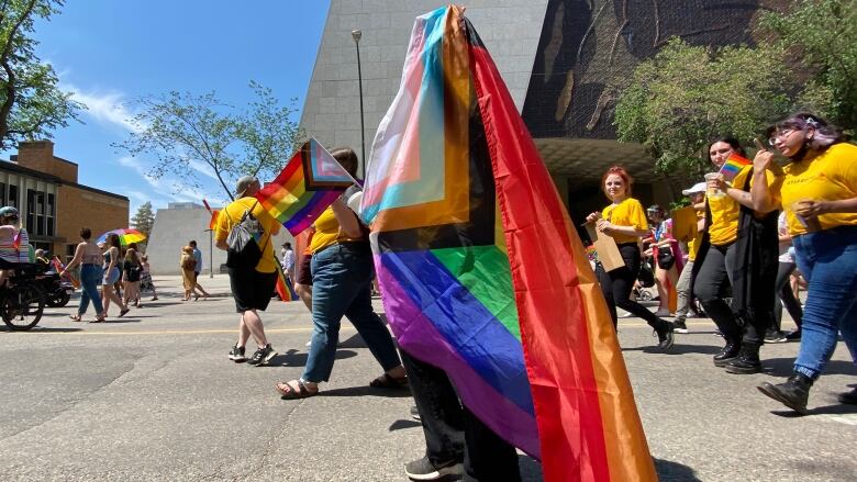 A colourful Pride flag is foreground, with people marching in yellow shirts behind.