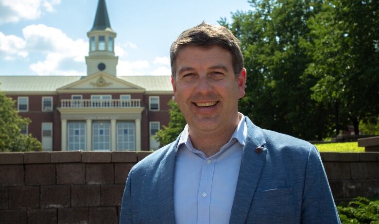 A smiling man in a blue dress shirt and blue blazer standing in front of a brick ledge with a building in the background