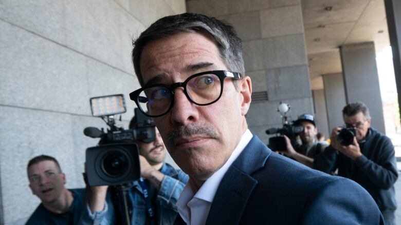  A man wearing glasses walks past reporters and cameras gathered at the entrance to the Montreal courthouse.