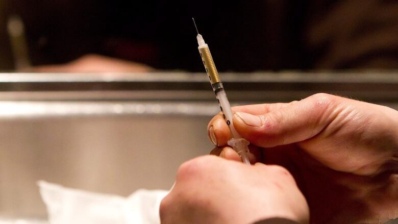 A man's hands holding a syringe are seen foreground, with his blurred reflection seen in a mirror in the background.