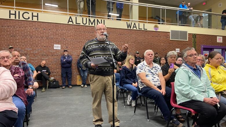 A many stands at a microphone at an outside event in front of people in people in chairs.