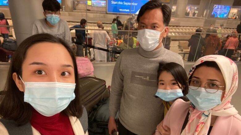 A woman wearing a face mask takes a selfie at an airport. She stands next to a man, a girl and a woman. 