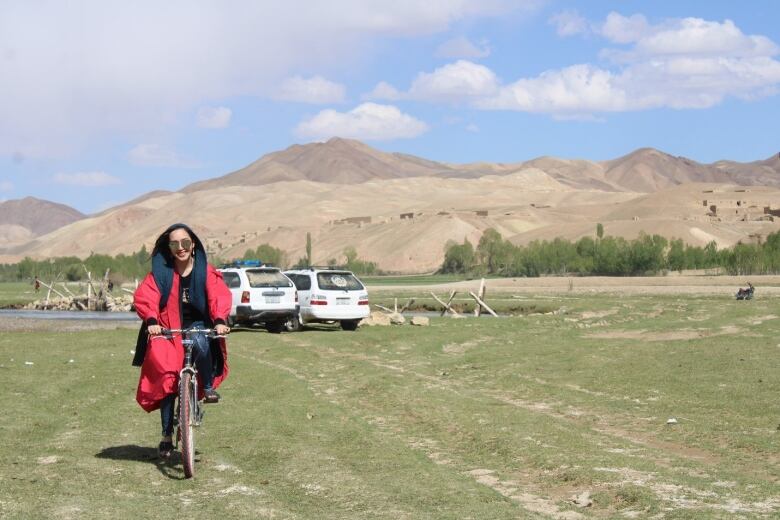 A woman rides a bike on a field with mountains in the distance.