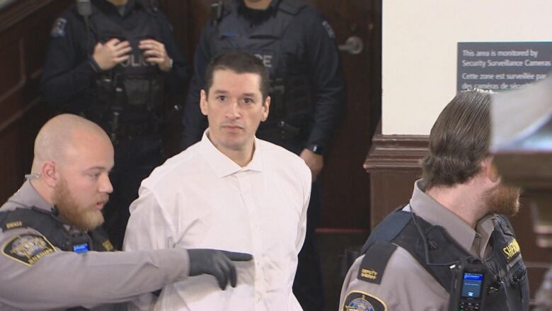 A middle-aged man in a white shirt and handcuffs is surrounded by sheriffs as he is led into a courtroom.