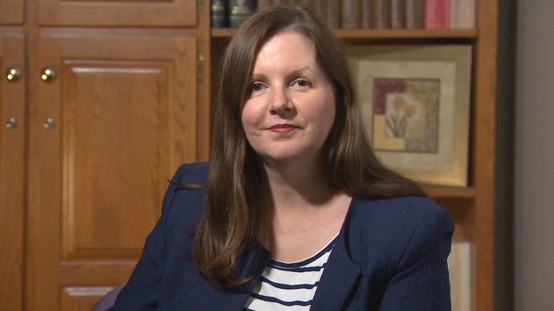 A portrait of a woman with long, dark hair, seated in what appears to be an office, wearing a navy blazer and navy and white striped shirt.