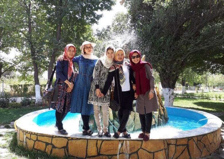 Five women link their arms around each other while standing in front of a fountain.