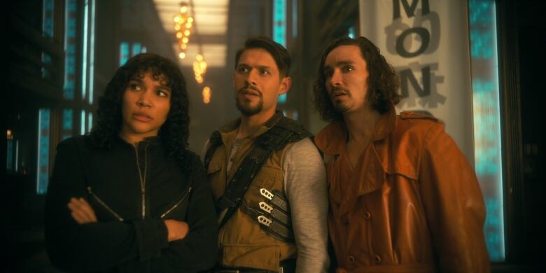 A woman with her arms crossed, a man with three throwing knives in a holster and another man in a leather jacket look off to the left with confused faces. 