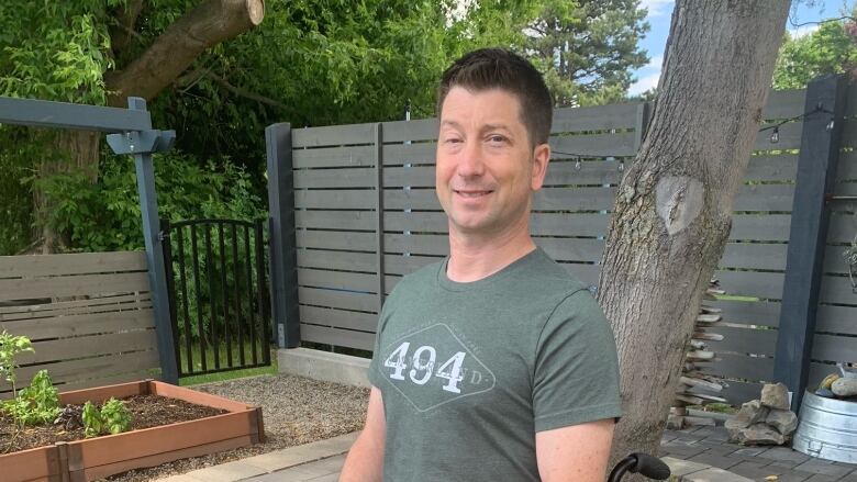 A man sitting in a wheelchair in an olive T-shirt in a fenced yard containing a tree and  plants in a planter smiles at the camera