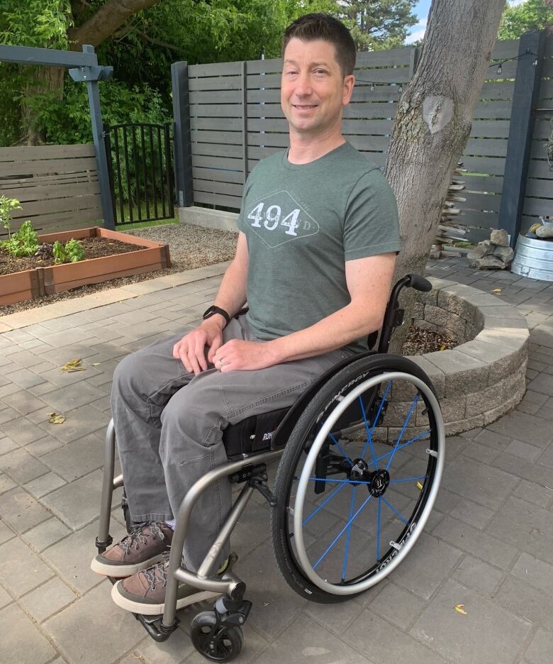 A man sitting in a wheelchair in an olive T-shirt in a fenced yard containing a tree and  plants in a planter smiles at the camera