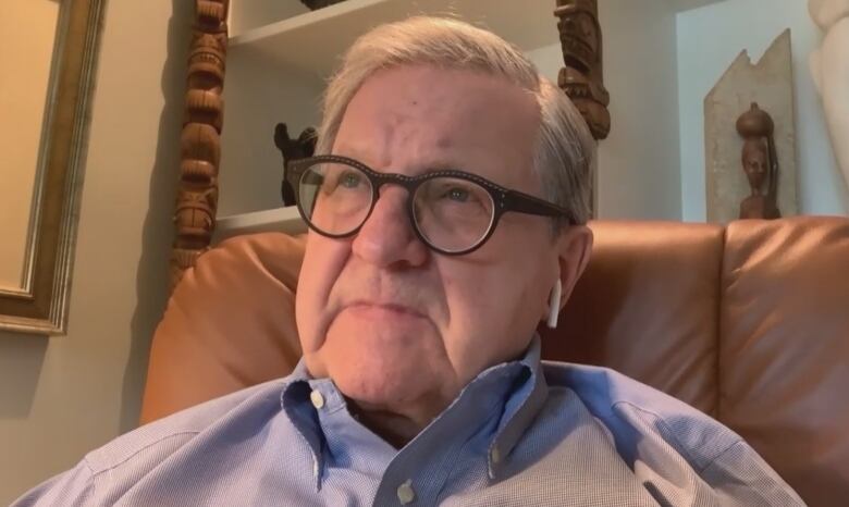 A grey-haired man wearing glasses sits in a brown desk chair in front of shelves lined with art pieces.