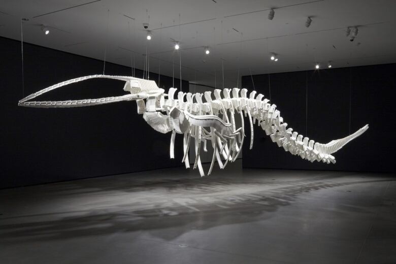 Photo of Cetology by Brian Jungen, a whale skeleton made of white plastic lawn chairs, which has been suspended from the ceiling in a dark gallery.