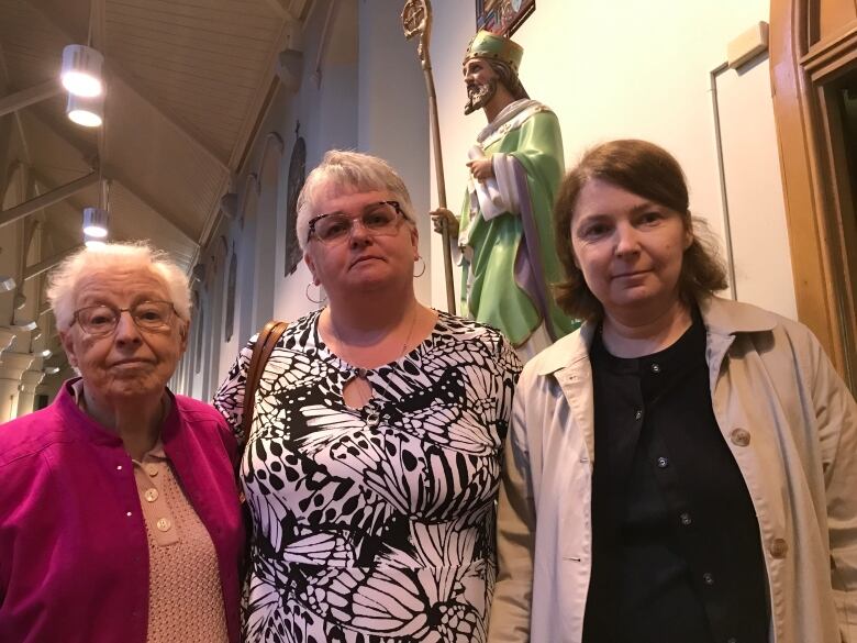 Three women stand side by side in a church in St. John's. a large statue depicting a man with a ctoss stands behind them.