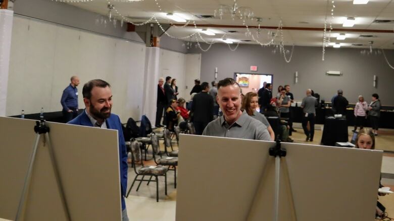 Two men look at a display board in a meeting room, with other people in the background.