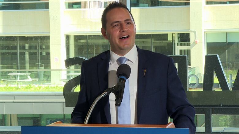 A man in a blue suit is seen talking with the help of a mic.