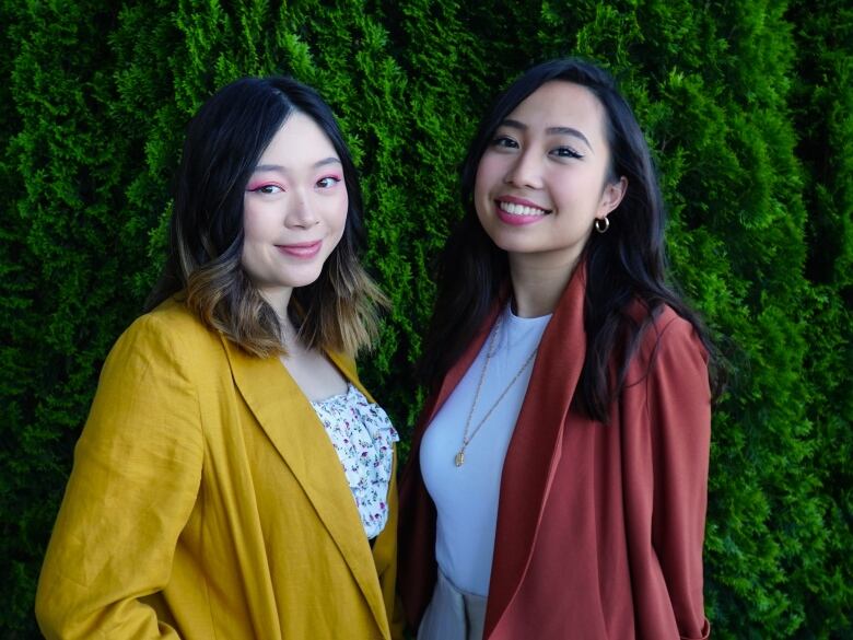 Two women, one in yellow and another in rusty orange, smile at the camera. There is a large bush behind them.