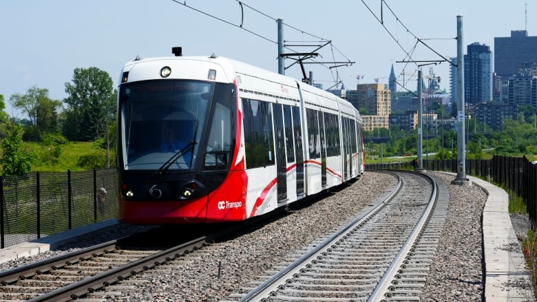 A red and white light rail train on the tracks with the Ottawa skyline in the background.