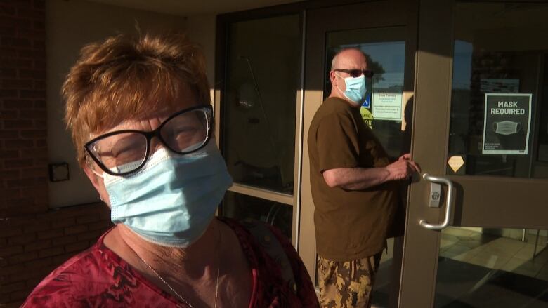 A woman wearing a face mask outside a building, with a man wearing a face mask opening door to building behind her.