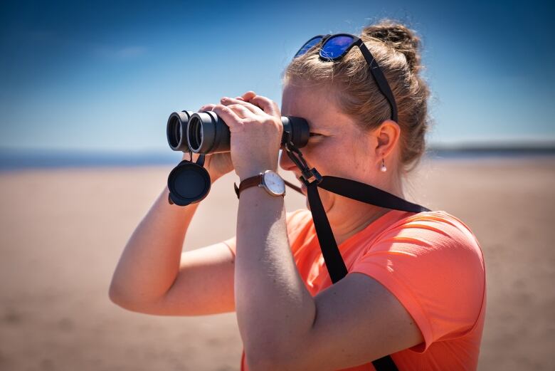 Woman with sunglasses on top of head looks through binoculars at a beach.