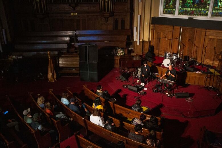 Aerial view of a stage with two performers dressed in all black. one playing a keyboard and another holding a microphone while a crowd seated in pews watch