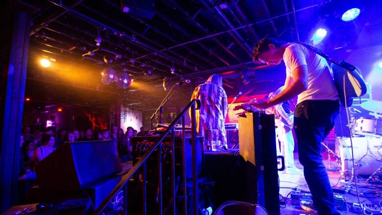 A man in a white shier plays a musical instrument on stage as a crowd looks on. The area is covered in coloured lights