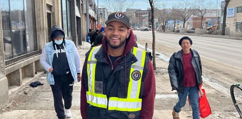 A man in a high vis vest and ball cap stands on a sidewalk as pedestrians pass by.