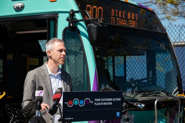 A white-haired man in a grey blazer stands in front of a podium near a purple and yellow bus. The podium reads 'Your Customer action plan in motion'.