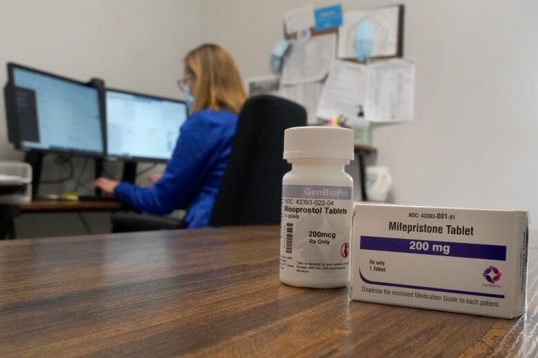 A box of Mifepristone tablets is in the foreground. A woman working at a computer in the background.