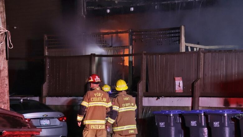 Two firefighters, wearing brown uniforms, hold a hose and direct it at a fire in the background of the image. Their uniforms have bright yellow lettering that says 'Port Coquitlam'