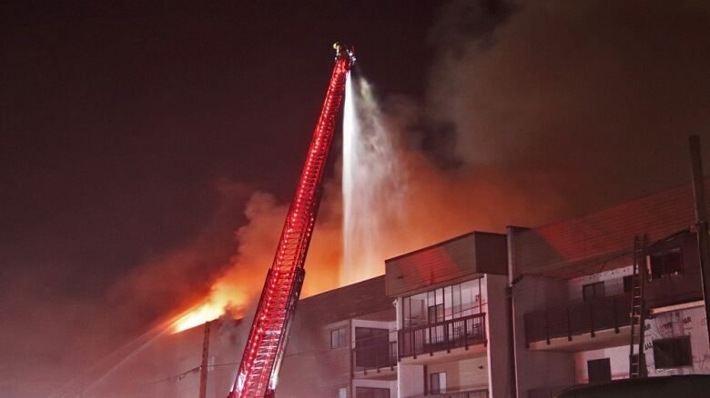 A firefighter stands on the end of a very long extended ladder and pours fire atop a building. Red lights and smoke are visible.