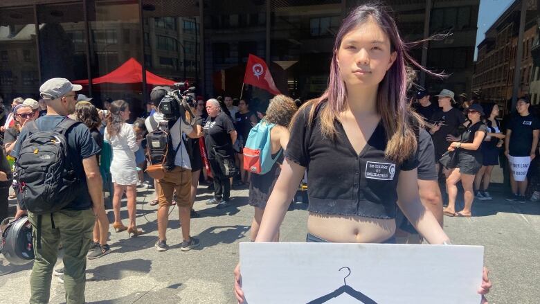 A person with long purple hair and a black t-shirt holds a wite sign with a coat hanger on it, that says, 'Never again.'