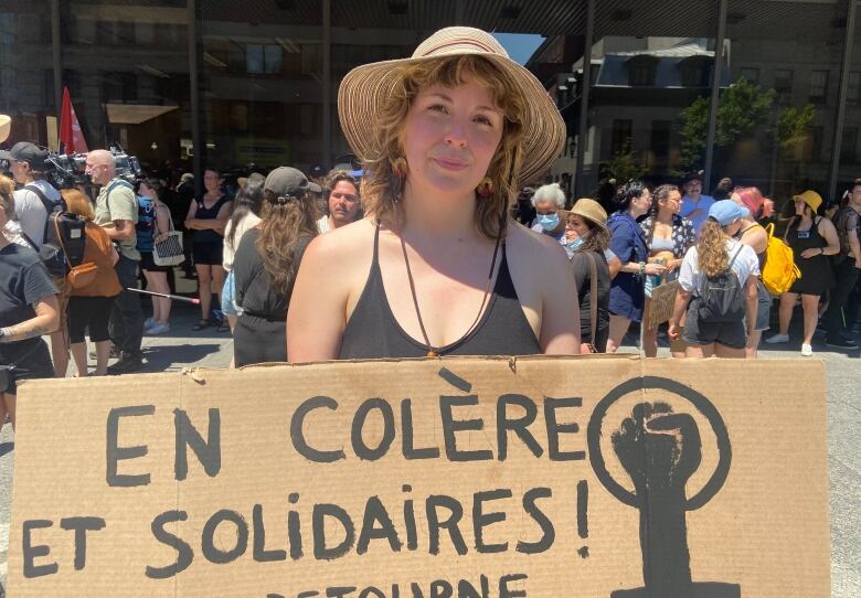 A blond woman with a straw hat holds up a cardboard sign that say, 'angry and in solidarity' with a fist painted in black and a circle around it. 