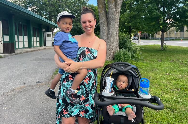 A mother stands smiling with her son in her arm and a younger child in a stroller. 