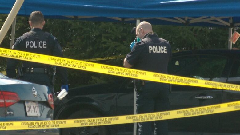 An RCMP member photographs the scene outside a Logan Lane duplex in Moncton's north end after 18-year-old Joedin Leger was fatally shot on April 25, 2022. A car is under a crime scene tent and yellow police tape is in the foreground. 