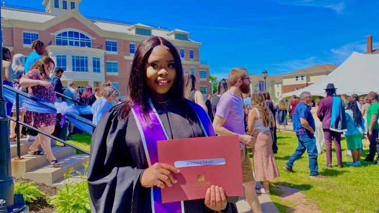 Young smiling woman wearing graduation gown and holding diploma stands in front of brick building. 