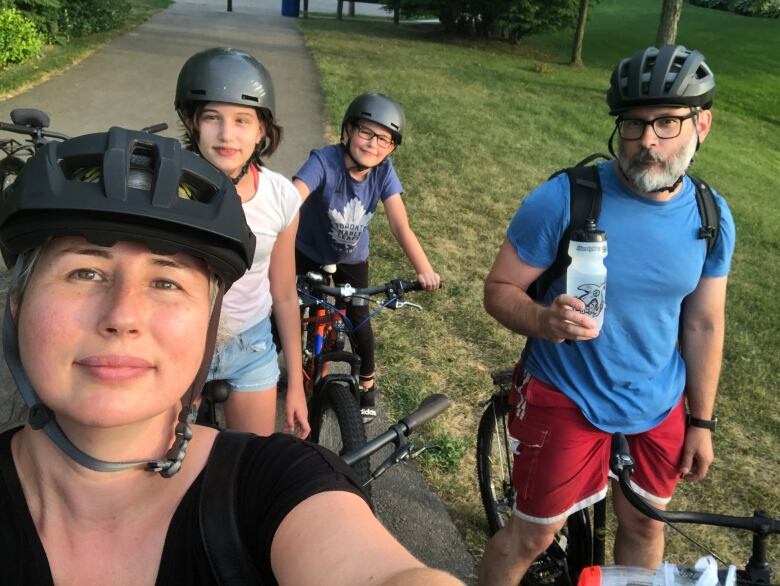 A woman wearing a bicycle helmet stands next to two children and one man all on bicycles. 