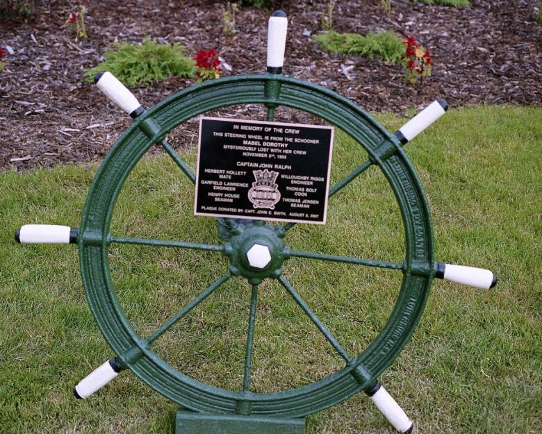 A green ship's steering wheel with white handles sits in a park. There is a plaque mounted near the top of the wheel.