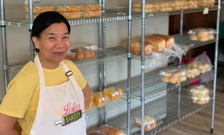 A woman wearing an apron reading 'Lola's Bakery' stands in front of shelves of baked goods.